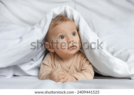 Cute little baby playing hiding under white blanket at home.