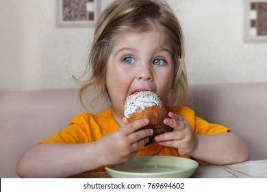 A cute little baby is sitting at a table eating a bun. Child with blue eyes