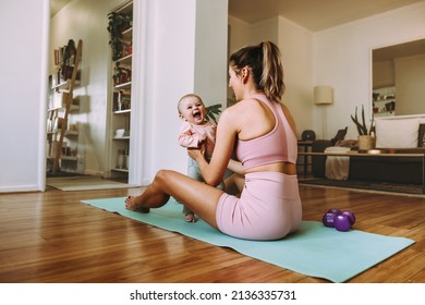 Cute Little Baby Smiling Excitedly In Her Mother's Hands. Happy Mom Working Out With Her Baby On An Exercise Mat At Home. New Mom Bonding With Her Baby During Her Post-natal Fitness Routine.