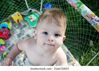 Cute Little Baby Who Can't Walk Yet Is In Play Pen. Naked Baby Stands In An Play Pen And Looks Up. Toddler Spending Time Outdoors On A Summer Day, Nature.