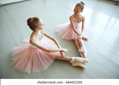 Cute Little Ballerinas In Pink Costume And Point Shoes Is Sitting On The Floor. Kids In Dance Class. Child Girl Is Studying Ballet In Pink Skirt. Two Little Ballet Girls Sit In 