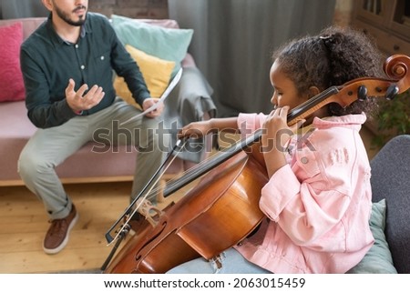 Cute little biracial girl gliding fiddlestick across cello strings while sitting in front of her music teacher
