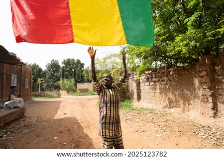 Cute little black African boy reaching out for the malian flag hanging over him on a rural village road