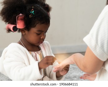 Cute Little Black African Girl Wearing White Robe, Playing Dress Up And Putting Nail Polish On Diverse Mom's Hand.