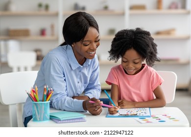 Cute Little Black Girl With Bushy Hair Exercising At Daycare, Sitting At Table With Friendly Female Teacher And Doing Various Tests, Young Woman Psychologist Working With Child Preschooler