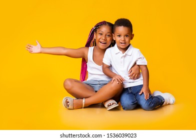 Cute little black girl cuddling her adorable baby brother, sitting on floor, looking aside and gesturing over yellow background, copy space - Powered by Shutterstock