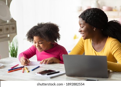 Cute Little Black Girl Drawing In Kitchen Next To Her Mom Working Online On Laptop. Young African American Mother Sitting At Table With Computer Spending Time With Her Kid And Enjoying Freelance Work