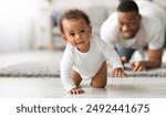 Cute Little Black Infant Baby Crawling On Floor At Home, Proud Young Father Looking At Him And Smiling, Dad And Toddler Child Enjoying Spending Time Together, Selective Focus With Copy Space