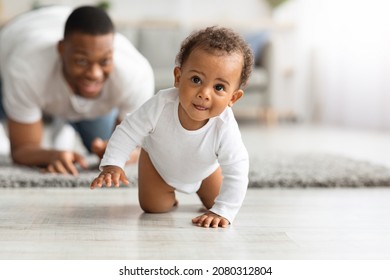 Cute Little Black Infant Baby Crawling On Floor At Home, Proud Young Father Looking At Him And Smiling, Dad And Toddler Child Enjoying Spending Time Together, Selective Focus With Copy Space