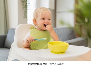 Cute little blond baby having lunch in green bib sitting at table with high chair holding spoon with meal in yellow plate in front of him, looking aside smiling, putting food in mouth with hand