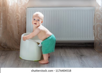 Cute little blond caucasian boy in funny green shorts playing with white potty indoor. Adorable child learning about potty-chair during game. Smiling positive kid with heating radiator on background