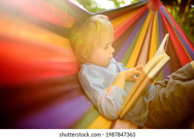 Kleiner blonder Kaukasier, der Buch liest und sich mit mehrfarbiger Hängematte im Hinterhof oder auf dem Spielplatz im Freien amüsiert. Sommeraktive Erholung für Kinder. Kinderschwingen und Entspannen in der Hängematte.