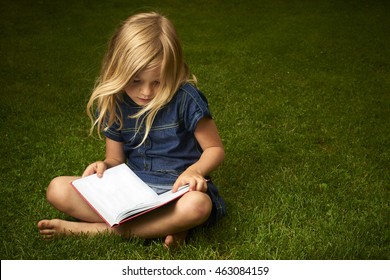 Cute Little Blond Girl Reading Book Outside On Grass