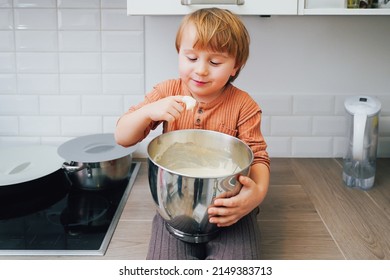Cute Little Blond Preschool Kid Boy Baking Cake In Domestic Kitchen, Indoors. Smiling Child  Tasting Sweet Dough. Little Helper