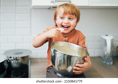 Cute Little Blond Preschool Kid Boy Baking Cake In Domestic Kitchen, Indoors. Laughing Child  Tasting Sweet Dough. Little Helper