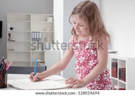 Cute little blonde girl in a pretty floral summer dress smiling happily as she stands at a table sketching on a large sketchpad