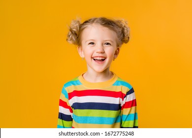 Cute little blonde girl smiling holding hands on the sides stands on a yellow background. a happy child in a multicolored striped jacket on a yellow background. space for text