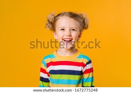 Cute little blonde girl smiling holding hands on the sides stands on a yellow background. a happy child in a multicolored striped jacket on a yellow background. space for text