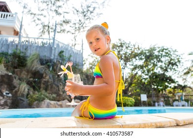 Cute little blonde girl swimming in big pool. She sit on the pool edge. Little lady hold big glass with water, straw and frangipani and look to the camera. Sunbathing and leisure on sunny summer day.