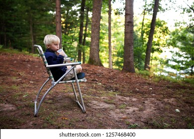 A Cute Little Blonde Kid Is Drinking Juice As He Sits In A Vintage Lawn Chair While Camping Near The Lake.