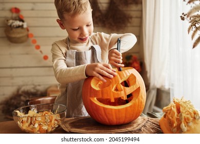 Cute little boy in apron scooping out all pulp from large orange pumpkin while carving classic traditional jack-o-lantern with scary face for Halloween celebration with family
