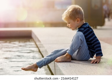 A Cute Little Boy, Barefoot, Wants To Climb Into The Water At The City Fountain During The Summer.