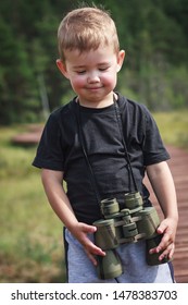 Cute Little Boy In A Black T-shirt Standing With Binoculars Around His Neck And Smiling Embarassed At Outdoor Nature