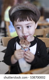 Cute Little Boy In A Brown Newsboy Cap And Tuxedo T-shirt Eating A Messy Chocolate Ice Cream Cone