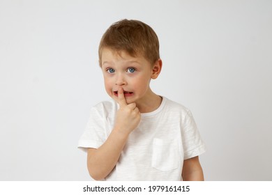 Cute Little Boy In Casual Clothing Standing On White Background Of Studio And Putting Index Finger On Lips While Looking At Camera. The Child Keeps A Secret