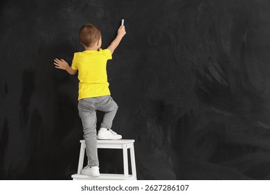 Niño lindo con el dibujo de la tiza en la pared negra, vista posterior