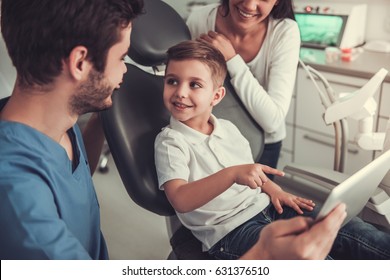 Cute Little Boy At The Dentist. Mom Is Supporting Her Son And Doctor Are Using A Digital Tablet