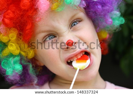A cute little boy dressed up in a clown costume, making a silly face and eating a lollipop