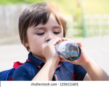 Cute Little Boy Drinking Colding From Clear Glass Bottle In The Hot Sunny Day On Summer, Kid Boy Drink Soda Or Soft Drink Sitting In Street With Blurry Background