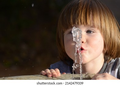 Cute Little Boy Drinking From A Fountain. Summer Thirst. A Baby With Long Hair Catches A Jet Of Water With His Mouth. Drinking Fountain At The Playground