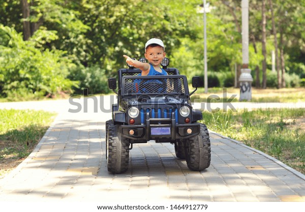 A cute, little boy drives an electric black car\
in a summer park.	