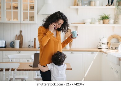 Cute Little Boy Embraces His Mom  At Kitchen While She Is Talking By Phone Holding Cup, Smiling, Looking At Son. Cheerful Hispanic Young Woman In Orange  Blouse Happy At Home With Beloved Little Boy.