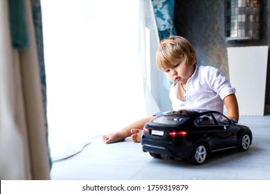 A cute little boy of European appearance is sitting on the floor of the house and playing with a big black toy car. The concept of child development at home. Focus on the child.