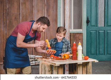 Cute Little Boy With Father In Apron Preparing Hot Dog Together In Backyard, Dad And Son Cooking Concept