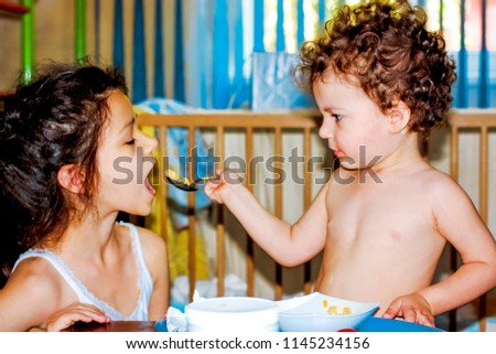 Cute little boy feeding baby sister in a her play room. Kids playing with toy dishes and kitchen. Little boy at lunch or breakfast with his big sister. Nutrition for children.
