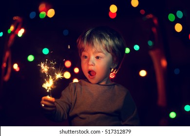 Cute Little Boy With A Funny Face Holding Sparklers. There Is A Background With Multi Colored Lights. Image With Selective Focus And Toning