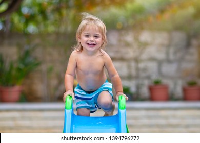 Cute Little Boy Having Fun In Backyard, With Pleasure Riding A Water Slide On The Beach Resort, Happy Child Enjoying Summer Holidays