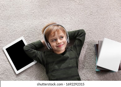 Cute little boy with headphones and tablet listening to audiobook on floor indoors, flat lay