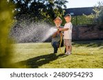Cute little boy and his grandfather watering lawn in backyard with gardening hose.