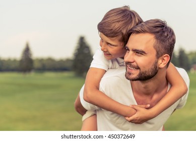 Cute Little Boy And His Handsome Young Dad Are Looking Forward And Smiling While Resting In The Park. Son Is Sitting Pickaback