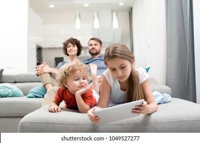 Cute little boy and his pretty elder sister lying on sofa and playing game on digital tablet while his parents sitting behind them and watching TV. - Powered by Shutterstock