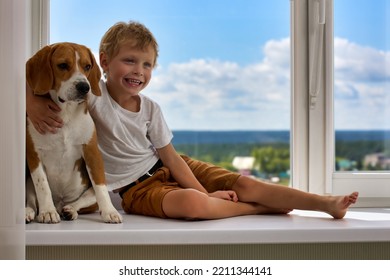 Cute Little Boy And His Thoroughbred Red Dog In Hug Sit On Wide Windowsill. Happy Child Hugs His Beloved Dog Sitting At Large Window Of Multi-storey Building