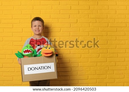 Cute little boy holding donation box with soft toys near yellow brick wall, space for text