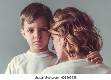 Cute Little Boy Is Hugging His Mom And Looking Sadly At Camera, On Gray Background