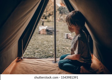 A Cute Little Boy Inside An Old Camping Tent Is Fulfilling A Metal Mug With A Rain Water. Image With Selective Focus, Noise Effect And Toning