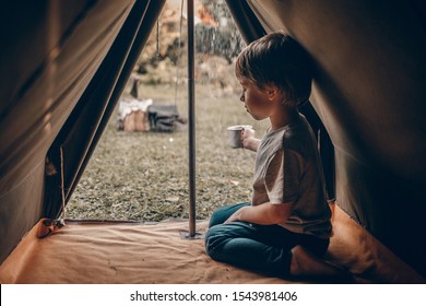 A Cute Little Boy Inside An Old Camping Tent Is Fulfilling A Metal Mug With A Rain Water. Image With Selective Focus, Noise Effect And Toning